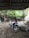 A motorbike is parked under a corrugated metal roof. The background includes a basketball hoop attached to a tree and lush green foliage. The ground appears to be dusty with some scattered leaves.