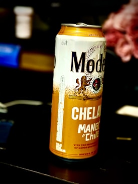 A can of Modelo Chelada with mango and chili flavor is prominently displayed. The can features a yellow and white color scheme with bold lettering and decorative elements.