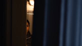 A dimly lit scene with a person partially visible through a narrow opening, suggesting subtle observation or secrecy, as if they are peering out from behind a door or curtain.
