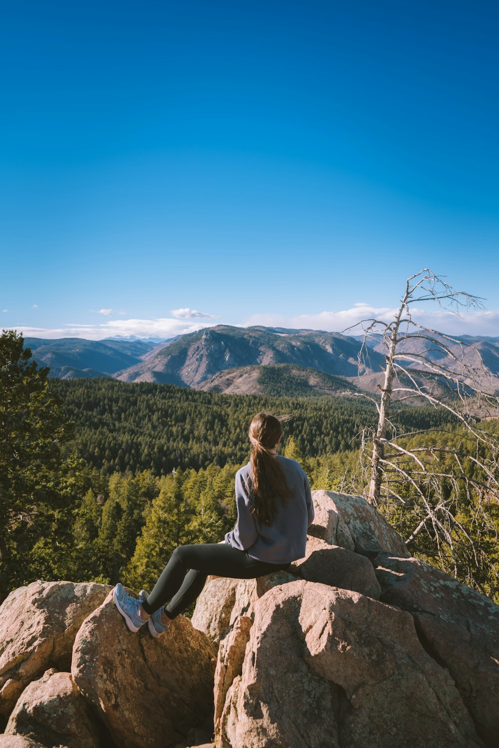 woman in white long sleeve shirt sitting on rock near green trees and mountains during daytime