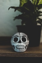 A decorative skull with intricate black designs is placed on a wooden surface. Behind it, a potted plant is slightly out of focus, adding depth to the composition.