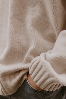 A close-up view of a person wearing a beige knitted sweater with ribbed cuffs. The lower edge of the sweater slightly overlaps a pair of blue jeans, where a hand is placed casually inside the pocket.