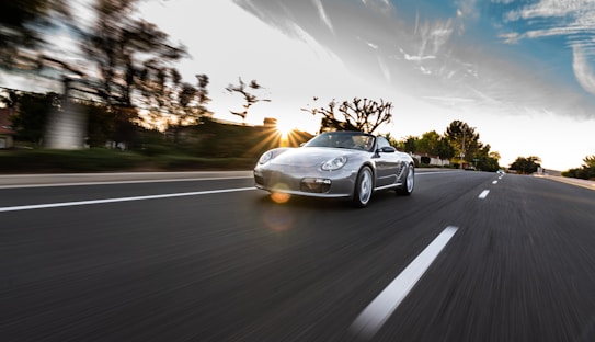 A sports car is driving on an open road with trees and buildings blurred in the background. The sun is setting, casting a warm glow on the scene with a blue and orange sky. The road is empty, adding a sense of speed and motion to the image.