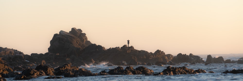 silhouette of person standing on rock formation in front of body of water during daytime