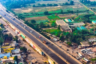 An aerial view of a busy highway running through an urban area with buildings, trees, and agricultural fields. Vehicles including cars, buses, and motorcycles are visible on the road. The urban landscape contains a mix of residential and commercial buildings, some with colorful roofs. Green fields and patches are evident in the background, suggesting agricultural activity.