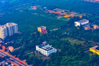 Aerial view of a semi-urban area featuring several buildings surrounded by lush greenery and agricultural fields. The landscape contrasts between developed infrastructure and vast green spaces, with roads weaving through the area.