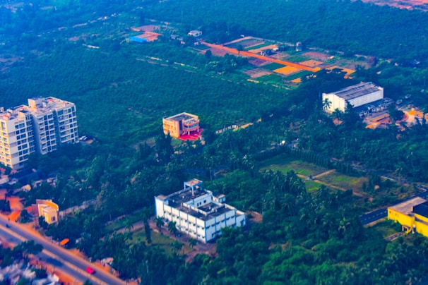Aerial view of a semi-urban area featuring several buildings surrounded by lush greenery and agricultural fields. The landscape contrasts between developed infrastructure and vast green spaces, with roads weaving through the area.