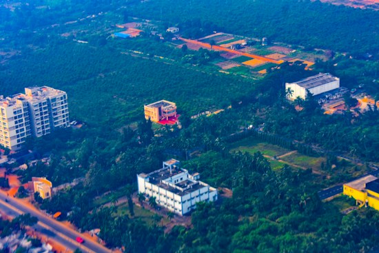 Aerial view of a semi-urban area featuring several buildings surrounded by lush greenery and agricultural fields. The landscape contrasts between developed infrastructure and vast green spaces, with roads weaving through the area.