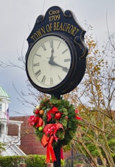 A large, freestanding outdoor clock displays Roman numerals and is labeled 'TOWN OF BEAUFORT' with 'Circa 1709' above it. A festive wreath with red ribbons and ornaments decorates the base of the clock. The background includes leafless trees and a building partially visible with an American flag and a porch.