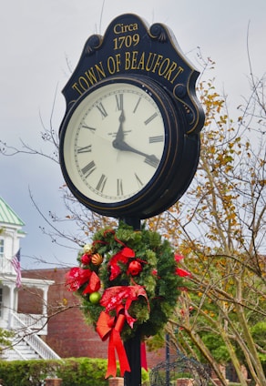 A large, freestanding outdoor clock displays Roman numerals and is labeled 'TOWN OF BEAUFORT' with 'Circa 1709' above it. A festive wreath with red ribbons and ornaments decorates the base of the clock. The background includes leafless trees and a building partially visible with an American flag and a porch.