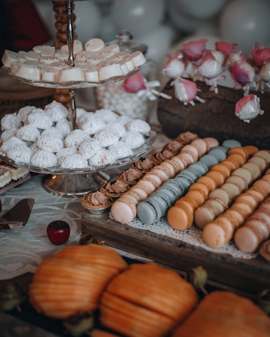 An assortment of desserts is displayed on a table with various treats including powdered cookies, macarons in different colors, and decorated cake pops. The scene includes a multi-tiered stand holding some of the pastries and a lace tablecloth underneath the spread. There are also artificial pink flowers used as decor placed among the sweets.