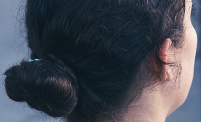 The back of a person's head is shown, with dark brown hair tied up in a bun. The hair appears slightly wavy and is secured with a small hairband. The image captures the side profile of the head, focusing on the ear and the texture of the hair.