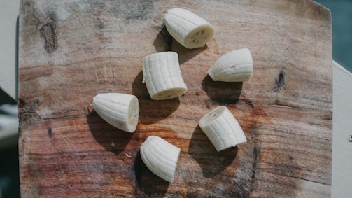 Several pieces of a peeled banana are scattered on a wooden cutting board. The board has a textured, rustic appearance with visible grain patterns and knots.