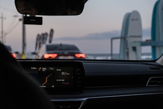 The interior of a car with a digital display showing various metrics is visible. The view is looking out the windshield toward another car stopped ahead, identifiable by its illuminated tail lights against the evening sky. Decorative flags and a structure are visible on the roadside.