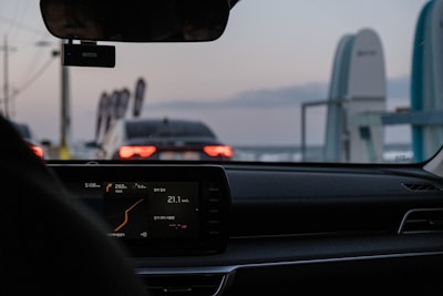 The interior of a car with a digital display showing various metrics is visible. The view is looking out the windshield toward another car stopped ahead, identifiable by its illuminated tail lights against the evening sky. Decorative flags and a structure are visible on the roadside.