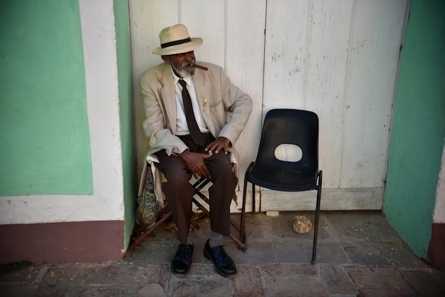 An elderly man with a white beard is sitting on a folding chair against an aged wooden wall. He wears a light-colored suit and a straw hat, with a cigar in his mouth. Next to him is an empty black plastic chair. The ground is stone, and the wall has green accents.