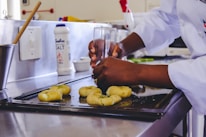 A person wearing a white chef's coat is shaping some dough with their hands using a glass, on a metal baking sheet in a kitchen. There is a container of salt and various kitchen utensils visible in the background.