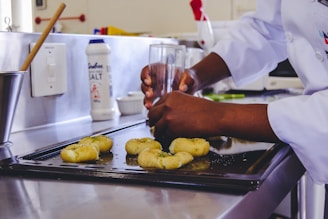 A person wearing a white chef's coat is shaping some dough with their hands using a glass, on a metal baking sheet in a kitchen. There is a container of salt and various kitchen utensils visible in the background.