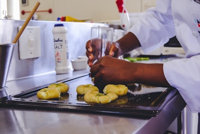 A person wearing a white chef's coat is shaping some dough with their hands using a glass, on a metal baking sheet in a kitchen. There is a container of salt and various kitchen utensils visible in the background.