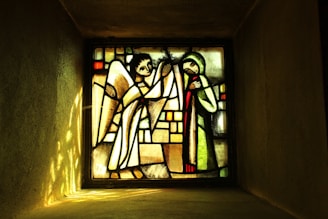 A stained glass window featuring an angel on the left with wings and a halo, and a figure on the right in a robe with hands clasped. The background is composed of geometric patterns in muted colors.