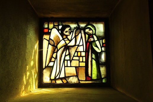 A stained glass window featuring an angel on the left with wings and a halo, and a figure on the right in a robe with hands clasped. The background is composed of geometric patterns in muted colors.