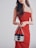 A woman wearing a strapless red dress is holding a small Prada handbag with metal chains. Her long, wavy brown hair is loose, and she stands against a plain, light background.