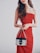 A woman wearing a strapless red dress is holding a small Prada handbag with metal chains. Her long, wavy brown hair is loose, and she stands against a plain, light background.