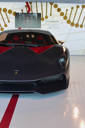 A sleek, black sports car with red accents is prominently displayed indoors. The vehicle has a low profile and aggressive styling. Behind the car, there is a decorative wall featuring geometric patterns and a screen. The words '6&deg; Elemento' are visible on the wall, suggesting a focus on advanced materials or technology.