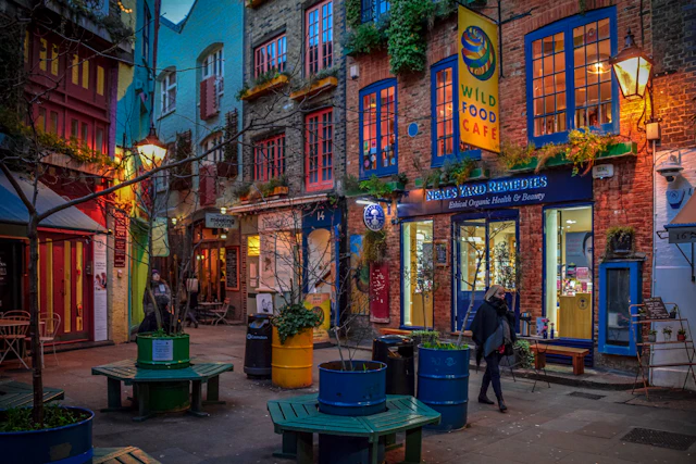 The colourful shopfronts and cafe of Neal's Yard in Covent Garden at dusk, with string lights and a person walking through.