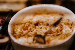 A close-up of a baked dish in a white round casserole dish, featuring a golden-brown, crispy top with hints of cheese and scattered breadcrumbs. A serving spoon is partially visible in the foreground, suggesting the dish is ready to be served.