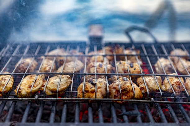 Chicken pieces are being grilled over charcoal on a metal grill grate. The chicken is lightly charred and appears to be marinated. Smoke rises from the grill, suggesting a lively cooking process. The background is slightly blurred, emphasizing the focus on the grilling.