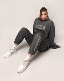 A person is sitting on the floor wearing a dark grey hoodie and matching jogger pants. The hoodie has the word 'HIELO' printed on it. They are also wearing white athletic shoes with Nike socks. The individual has long hair styled in a ponytail and is looking directly at the camera.