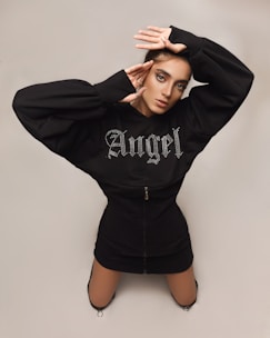 A person is posing against a plain background. They are wearing a stylish black outfit with the word 'Angel' adorned in rhinestones on the top. The individual is posed with both hands framing their face, displaying elaborate eye makeup and an intense gaze.