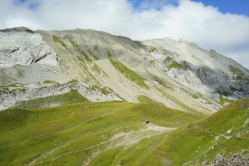 A vast and rugged landscape featuring large gray rock formations and expansive green hills. The terrain appears steep and rocky with patches of grass. A small path winds through the hills, suggesting possible hiking activity. The sky is partly cloudy, casting soft shadows over the scenery.