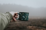 A hand holding a warm insulated mug outdoors with a blurred nature background.