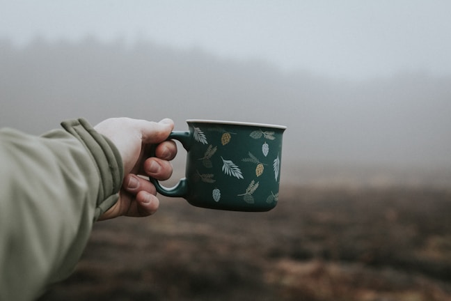 A hand holding a warm insulated mug outdoors with a blurred nature background.
