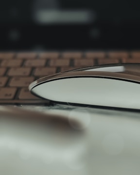 A computer mouse is placed on a reflective surface in front of a blurred keyboard. The focus is on the sleek lines and curves of the mouse, creating a modern and minimalistic aesthetic.