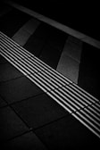 A monochromatic image of a tiled surface with a tactile paving strip featuring raised lines perpendicular to the direction of the tiles. The tiles vary in shades of grey and black, creating a geometric pattern.