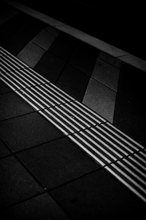 A monochromatic image of a tiled surface with a tactile paving strip featuring raised lines perpendicular to the direction of the tiles. The tiles vary in shades of grey and black, creating a geometric pattern.