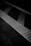 A monochromatic image of a tiled surface with a tactile paving strip featuring raised lines perpendicular to the direction of the tiles. The tiles vary in shades of grey and black, creating a geometric pattern.