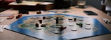A board game is laid out on a wooden table with hexagonal tiles and various wooden pieces, including houses and roads. Some tiles have numerical tokens on them. There is a box in the background, along with cards and a mobile phone nearby.
