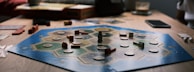 A board game is laid out on a wooden table with hexagonal tiles and various wooden pieces, including houses and roads. Some tiles have numerical tokens on them. There is a box in the background, along with cards and a mobile phone nearby.