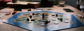 A board game is laid out on a wooden table with hexagonal tiles and various wooden pieces, including houses and roads. Some tiles have numerical tokens on them. There is a box in the background, along with cards and a mobile phone nearby.