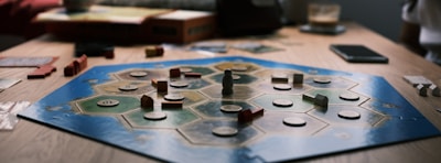 A board game is laid out on a wooden table with hexagonal tiles and various wooden pieces, including houses and roads. Some tiles have numerical tokens on them. There is a box in the background, along with cards and a mobile phone nearby.