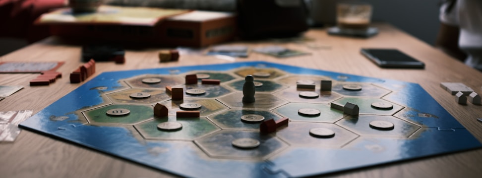 A board game is laid out on a wooden table with hexagonal tiles and various wooden pieces, including houses and roads. Some tiles have numerical tokens on them. There is a box in the background, along with cards and a mobile phone nearby.