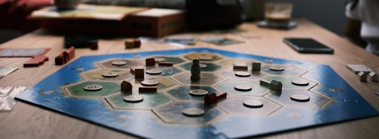 A board game is laid out on a wooden table with hexagonal tiles and various wooden pieces, including houses and roads. Some tiles have numerical tokens on them. There is a box in the background, along with cards and a mobile phone nearby.