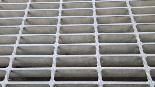 A close-up view of a metal grid or grating with evenly spaced horizontal and vertical bars. The bars intersect at right angles, forming a pattern of rectangular openings.