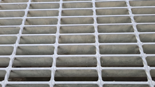 A close-up view of a metal grid or grating with evenly spaced horizontal and vertical bars. The bars intersect at right angles, forming a pattern of rectangular openings.