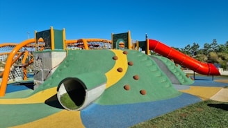A colorful playground structure featuring a green and yellow climbing mound with brown dome-shaped grips. A large, red curvy slide extends from the top of the mound. Orange arches form a climbing net above the structure, and the surrounding area is made of a soft, multicolored surface. Trees and a clear blue sky are in the background.