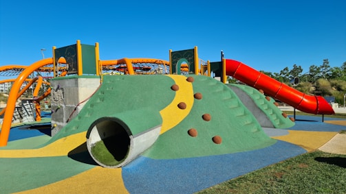 A colorful playground structure featuring a green and yellow climbing mound with brown dome-shaped grips. A large, red curvy slide extends from the top of the mound. Orange arches form a climbing net above the structure, and the surrounding area is made of a soft, multicolored surface. Trees and a clear blue sky are in the background.